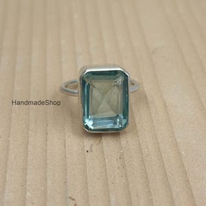 Peut inclure: Une bague en argent avec une grosse pierre précieuse rectangulaire bleu clair. La pierre est sertie dans une lunette en argent et a un aspect facetté. La bague est présentée sur une surface marron clair. Le texte "HandmadeShop" est visible.