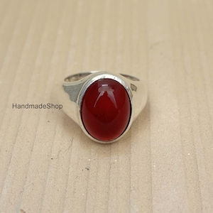 Puede incluir: Un anillo de plata con una gema grande, ovalada y de color rojo intenso. La gema está engastada en una banda de plata pulida. El anillo se muestra sobre una superficie neutra y texturizada. El texto "HandmadeShop" es visible.