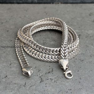 Può includere: Una collana a catena color argento con un design intrecciato, appoggiata su una superficie grigia. La collana ha una chiusura a moschettone. Il testo "HandmadeArtShop" è visibile.