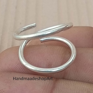 May include: A silver-colored ring with a unique, open design. The ring features a swirling, overlapping pattern, with a smooth, polished finish. The ring is shown on a light brown surface. The text "HandmaadeshopArt" is visible.