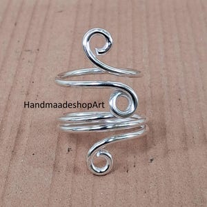 May include: A silver ring with a unique spiral design. The ring has a coiled band that wraps around the finger, with decorative swirls at the top and bottom. The ring is shown on a light brown surface.