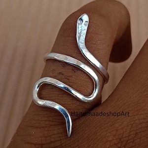 Puede incluir: Un anillo plateado con forma de serpiente que se enrolla alrededor de un dedo. El diseño del anillo presenta una cabeza y un cuerpo de serpiente, con un acabado texturizado. La cabeza de la serpiente tiene dos pequeñas hendiduras para los ojos. Es una joya.