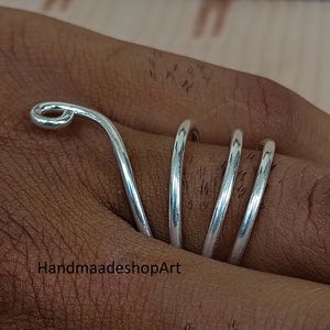 May include: A silver ring with a unique spiral design. The ring wraps around the finger with a curved, looped top. The ring is on a brown skin tone. The text "HandmaadeshopArt" is at the bottom of the image.