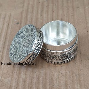 May include: A small, round, silver-colored metal container with a removable lid. Both the lid and the side of the base feature intricate, embossed floral and scrollwork patterns. The interior of the container is smooth and polished silver-colored metal.