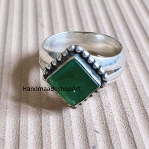 May include: A silver ring with a square, emerald-green gemstone. The gemstone is set in a bezel with a beaded border. The ring band is thick and has a split design. The text "HandmaadeshopArt" is visible.