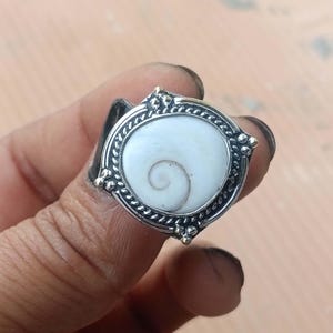 Puede incluir: Un anillo de plata con una incrustación circular de concha de ojo de Shiva blanca con un patrón de espiral natural. El engaste está enmarcado por un borde de cuerda trenzada y acentuado con pequeñas cuentas de plata en las cuatro esquinas del soporte.