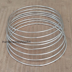 May include: A stack of seven thin, circular silver-colored metal bangles arranged in a conical formation. The smooth, reflective rings are layered one on top of the other, decreasing in diameter toward the top. The text HandmaadeshopArt is visible.