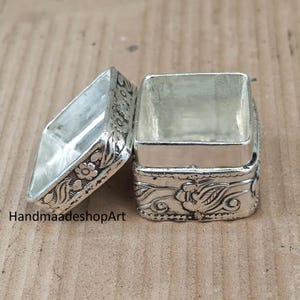 Può includere: Una piccola scatola rettangolare in metallo color argento con coperchio aperto. Le superfici esterne della scatola e del coperchio presentano motivi floreali e di foglie in rilievo. Il testo HandmaadeshopArt è visibile nell'angolo in basso a sinistra.