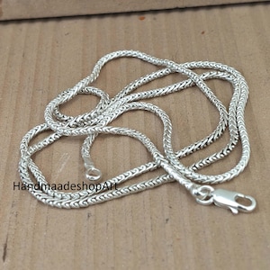 Puede incluir: Un collar de cadena plateada con un diseño tejido. El collar está sobre una superficie marrón. El cierre y el lazo final son visibles. El texto "HandmaadeshopArt" es visible en la parte inferior izquierda.