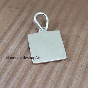 Puede incluir: Un colgante cuadrado de color plateado con una superficie lisa y plana y una anilla redondeada en la parte superior para su sujeción. El texto HandmaadeshopArt está impreso en negro en la parte inferior izquierda de la pieza metálica cuadrada.
