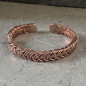 May include: A copper cuff bracelet with a woven design. The open-ended bracelet is made of interlocking copper links, creating a textured surface. The bracelet is displayed on a gray surface. The text "HandmadeArtShop" is visible on the left side.