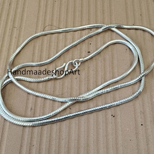 May include: A silver-colored snake chain necklace with a lobster clasp. The chain is made of small, interconnected links, creating a smooth, flexible design. The necklace is displayed on a light brown surface. The text "HandmaadeshopArt" is visible.