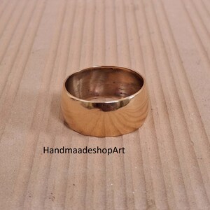 May include: A broad, gold-coloured ring with a smooth, polished surface. The ring has a rounded shape and a thick band. The text "HandmaadeshopArt" is visible on the surface below the ring.