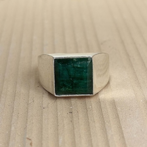 May include: A silver ring with a large, square-cut, deep green emerald gemstone. The ring has a simple, modern design with a polished finish. The emerald is set flush within the silver band, creating a sleek and elegant look.