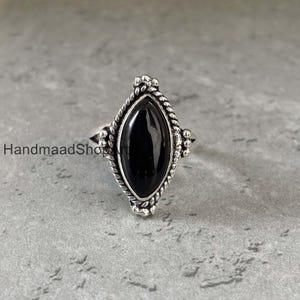 Puede incluir: Un anillo de plata con una gran piedra preciosa negra ovalada. La piedra preciosa está engastada en un marco de plata decorativo con un borde en forma de cuerda. El anillo tiene un estilo vintage, con una banda texturizada y detalles ornamentados.