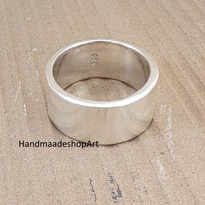 May include: A wide, polished silver ring with a smooth, flat surface. The ring has a simple, minimalist design and is marked with "925" on the inside. The ring is displayed on a light brown, textured surface.