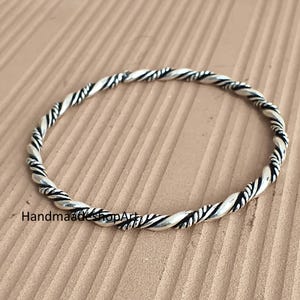 Könnte beinhalten: Ein silberfarbenes Armband im Stil eines gedrehten Seils. Das Armband hat ein geflochtenes Design mit abwechselnd hellen und dunklen Abschnitten. Der Text "HandmadeShopArt" ist unten links sichtbar.