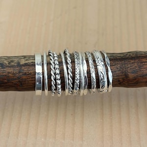 Puede incluir: Una colección de anillos de plata con varios diseños, incluyendo bandas lisas, patrones de cuerda trenzada y superficies texturizadas. Los anillos se muestran en una clavija de madera sobre un fondo neutro. Los anillos son de diferentes anchos y estilos.