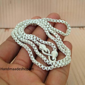 Puede incluir: Un collar de plata con un diseño de cadena de caja. El collar está hecho de un material metálico brillante y presenta un cierre seguro. La cadena se muestra sobre una superficie marrón claro.