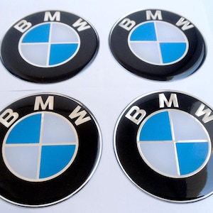 Op de afbeelding: Vier ronde BMW-emblemen. Elk embleem heeft een zwarte buitenring met zilveren letters "B", "M" en "W". Het midden heeft een blauw en wit kruisontwerp.