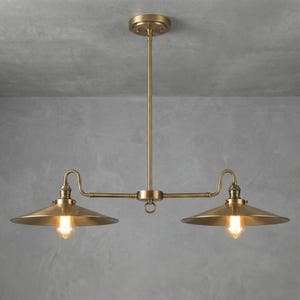 Op de afbeelding: Een messingkleurige hanglamp met twee lampen. De armatuur heeft een centrale staaf die aan een plafondbevestiging is bevestigd, met twee horizontale armen om de lampen vast te houden. Elke lamp heeft een conische kap en een zichtbare Edison-stijl lamp.