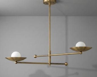 May include: A brass-toned, modern chandelier with two horizontal arms, each holding a white globe light within a shallow dish. The fixture has a central vertical rod and a ceiling mount.