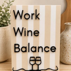 Könnte beinhalten: Ein rechteckiges Schild mit vertikalen beigen und weißen Streifen, das den schwarzen erhabenen Text Work, Wine, Balance sowie eine Grafik von zwei Weingläsern, die durch eine Wellenlinie verbunden sind, auf einer flachen Oberfläche zeigt.