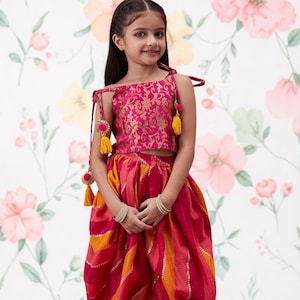 May include: A two-piece outfit consisting of a sleeveless hot pink crop top with gold floral patterns and tie-up shoulder straps with yellow tassels, paired with loose-fitting dhoti-style trousers featuring alternating diagonal stripes of pink and orange.