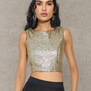 Könnte beinhalten: Ärmelloses, goldfarbenes Pailletten-Crop-Top mit schwarzem Besatz. Das Oberteil wird mit einer hoch taillierten schwarzen Hose kombiniert. Das Model trägt goldenen Schmuck, darunter ein Armband und Ohrringe. Der Hintergrund ist ein neutrales Grau.