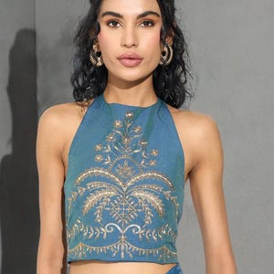 May include: A teal halter top with intricate gold embroidery. The design features a central floral motif with detailed patterns. The top is part of a set, paired with a matching blue tie-dye skirt. The style is suitable for special occasions.