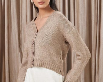 May include: A beige cardigan with a V-neck and button closure. The long-sleeved sweater is made of a knit material. The model is wearing the cardigan with white pants. The background is a beige curtain.
