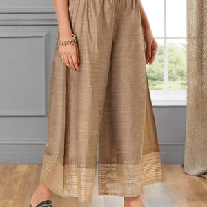 Può includere: Pantaloni palazzo beige a gamba larga e leggermente lucidi. L'orlo è decorato con un bordo a fantasia. La vita è elasticizzata. La modella indossa sandali neri.