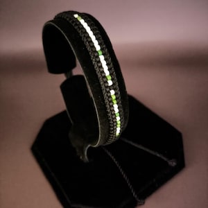 Peut inclure: Bracelet noir avec un motif tissé, orné de détails blancs et verts. Le bracelet est présenté sur un présentoir en velours noir, soulignant son design complexe et le contraste des couleurs. Un accessoire élégant.