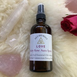 Può includere: Una bottiglia spray di vetro marrone con tappo nero ed etichetta bianca con la scritta "ALCHEMIST LOVE Ruby Rose Aura Spray". Cristalli e una rosa rossa sono disposti attorno alla bottiglia su una superficie bianca e soffice.