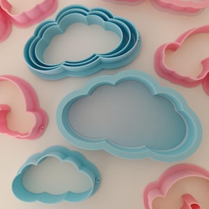 Può includere: Una collezione di formine per biscotti a forma di nuvola in varie dimensioni e colori. Le formine sono principalmente azzurre e rosa, con un semplice design a nuvola arrotondata. Le formine sono in plastica e sono disposte su una superficie bianca.