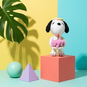 Op de afbeelding: Een gehaakte Snoopy-figuur met een roze verjaardagstaart met een brandende kaars. De figuur heeft zwarte oren en een gele kroon. Hij staat op een koraalkleurige kubus tegen een gele en blauwe achtergrond. Een groen blad en geometrische vormen zijn op de achtergrond.