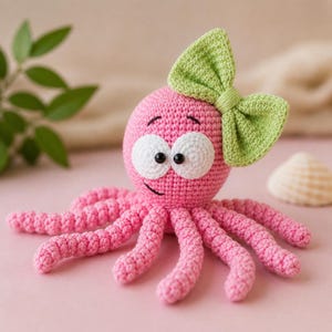 Pode incluir: Um polvo rosa feito à mão em crochê com um laço verde e olhos brancos grandes. O polvo tem oito tentáculos rosa e um rosto sorridente. Este brinquedo feito à mão é adequado para crianças.