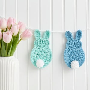 May include: Two crocheted bunny decorations in light blue and blue, each with a white pom-pom tail, hang from a white string. A vase of pink tulips sits to the left against a white background.