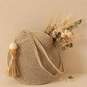 Puede incluir: Un bolso redondo tejido en beige con correas largas y un borla decorativa. La bolsa está llena de flores y follaje secos. La borla tiene un pompón color crema y una franja beige.