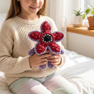 Op de afbeelding: Een handgemaakt Demogorgon-plush-speelgoed van gehaakte stof. De pluche heeft een rode, bloemachtige kop met witte stiksels en een zwart-wit midden. De bloemblaadjes zijn paars omlijnd. Het speelgoed wordt vastgehouden door een persoon die een crèmekleurige trui draagt.