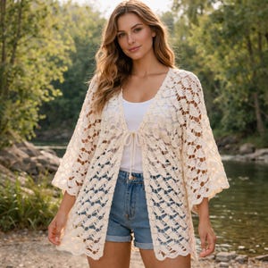May include: Cream-colored, open-front crochet cardigan with three-quarter sleeves. The cardigan features a tie closure at the waist and a delicate, openwork pattern. The model is wearing a white tank top and denim shorts.