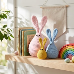 May include: A display of crochet bunnies in shades of pink, yellow, and blue, with black embroidered eyes. The bunnies are arranged on a wooden shelf alongside books, a wooden rainbow, and a blank banner.