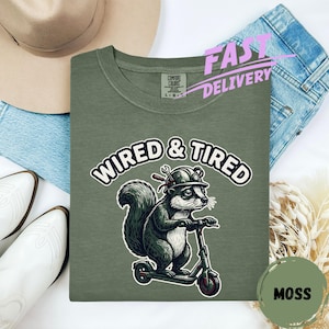 May include: Moss green Comfort Colors sweatshirt with a graphic of a squirrel riding a scooter, wearing a hard hat with tools, and the words "WIRED & TIRED". The shirt is laid flat with a hat, jeans, and boots in the background.