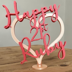 May include: A pink and gold cake topper with the words "Happy 21st Ruby" in a cursive font. The text is layered over a heart-shaped design. The topper is on a small, round base.