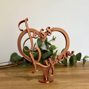 May include: A copper-coloured heart-shaped cake topper featuring the names "Jess" and "Ryan" in cursive script. The topper is mounted on a small, round base, ideal for wedding cakes or anniversary celebrations.