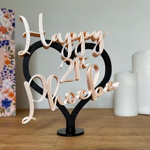 以下が含まれることがあります： 「Happy 21st Phoebe」の文字が筆記体で書かれたケーキトッパー。 ローズゴールドのグリッターテキストは、黒いハート型のフレームに配置されています。 トッパーは黒いベースに取り付けられており、21歳の誕生日に適しています。