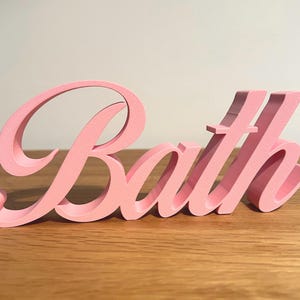 May include: A pink 'Bath' sign in a cursive font. The letters are in a rounded style, and the sign is placed on a wooden surface. The word is in a solid, three-dimensional form.