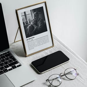 Op de afbeelding: Een zwart-witfoto in een lijst met de tekst "ya'aburnee" en een Arabische definitie. Een laptop, een smartphone en een ronde bril liggen ook op de tafel.