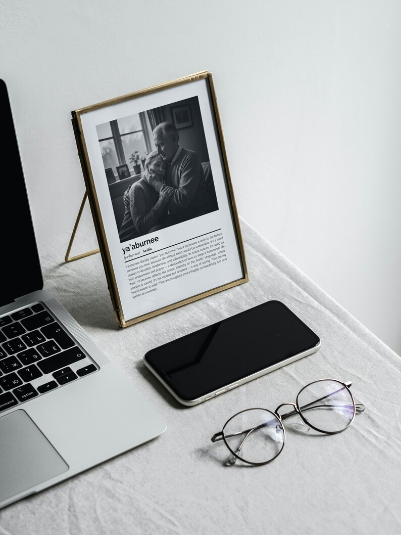 Op de afbeelding: Een zwart-witfoto in een lijst met de tekst "ya'aburnee" en een Arabische definitie. Een laptop, een smartphone en een ronde bril liggen ook op de tafel.