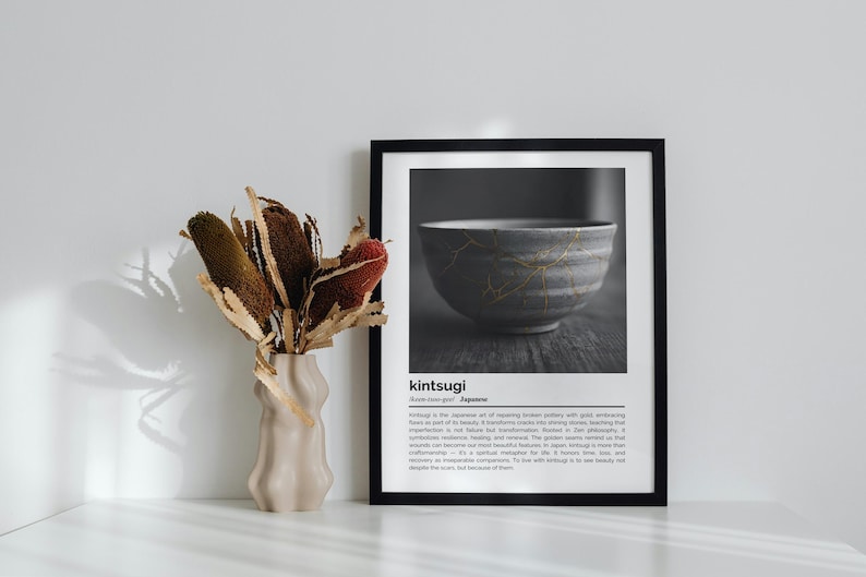 K&ouml;nnte beinhalten: Ein schwarz gerahmter Druck mit einem Graustufenbild einer japanischen Kintsugi-Schale mit goldenen Akzenten. Daneben steht eine beige Vase mit getrockneten Blumen. Der Text "kintsugi" ist sichtbar.